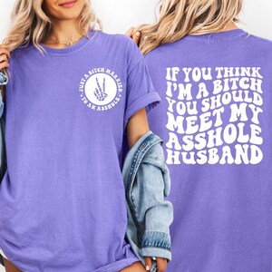 May include: A lilac-colored t-shirt with white text on the front and back. The front reads "Just a bitch married to an asshole" in a circle. The back says "If you think I'm a bitch you should meet my asshole husband."