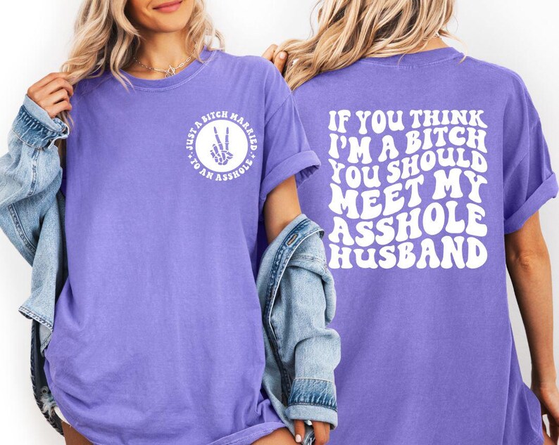 May include: A lilac-colored t-shirt with white text on the front and back. The front reads "Just a bitch married to an asshole" in a circle. The back says "If you think I'm a bitch you should meet my asshole husband."