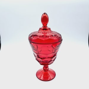 May include: A red glass candy dish with a lid. The dish has a pedestal base and a textured, faceted design.