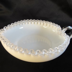 May include: A white milk glass bowl with a scalloped edge and a clear glass handle. The bowl has a silver-colored trim around the edge.