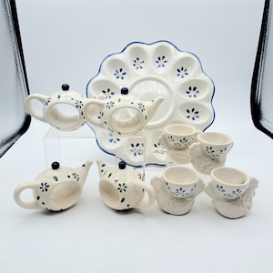 May include: A set of ceramic egg cups and napkin rings in a blue and white floral pattern. The egg cups are shaped like chickens and the napkin rings are shaped like teapots. There is also a matching egg plate with six individual egg cups.