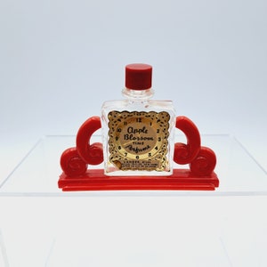 May include: A small, clear glass bottle of Apple Blossom Time Perfume with a red cap and a gold label. The bottle is sitting on a red plastic stand with a decorative design.