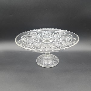 May include: A clear glass cake stand with a decorative, intricate pattern on the top plate and a pedestal base.