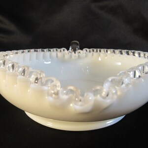 May include: White milk glass bowl with a silver trim and a ruffled edge. The bowl has a small handle on the top.