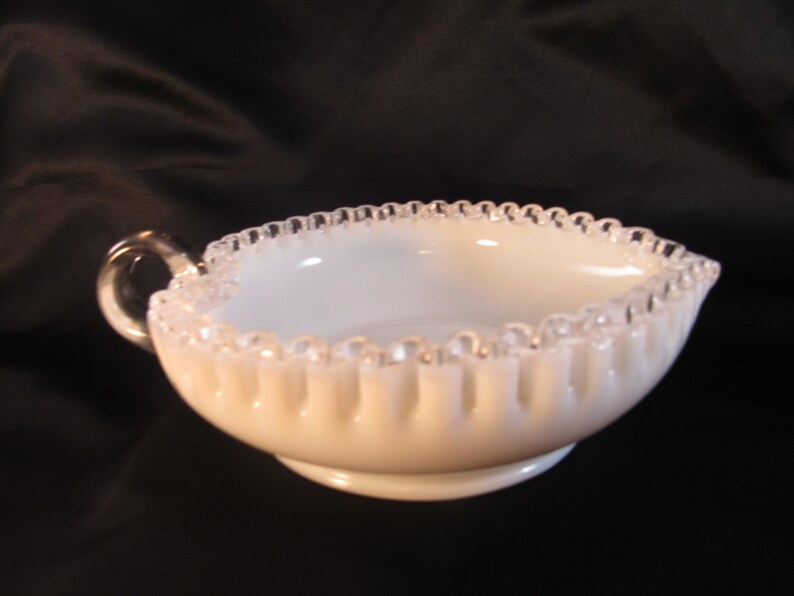 May include: A white milk glass creamer with a silver beaded edge and a handle. The creamer has a fluted design and a small base.