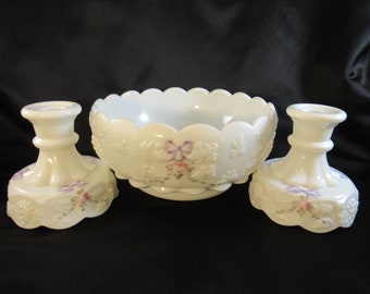 May include: A white milk glass bowl with a scalloped edge and a floral design. The bowl is part of a set with two matching candle holders. The candle holders have a scalloped base and a floral design with purple bows.
