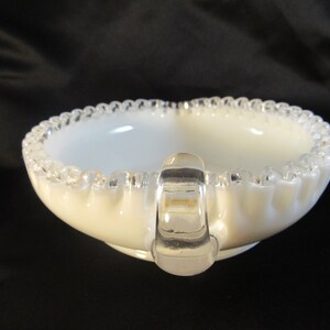 May include: A white milk glass bowl with a clear glass handle and a beaded rim. The bowl is a vintage style and has a scalloped edge.