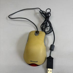 Puede incluir: Un ratón de ordenador Microsoft amarillo con un cable negro y un conector USB. El ratón tiene una luz roja en la parte inferior y el logotipo de Microsoft en el lateral.