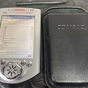 May include: A silver Compaq iPAQ pocket PC with a stylus and a black leather case. The screen displays a menu with various options. The case is embossed with the Compaq logo.