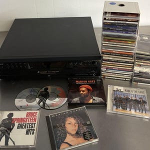 Puede incluir: Un reproductor de CD Sony negro con varios CD, incluidos álbumes de Eagles, Marvin Gaye, Bruce Springsteen, Mariah Carey y Kool & The Gang. Una pila de CD está en una caja de almacenamiento blanca.