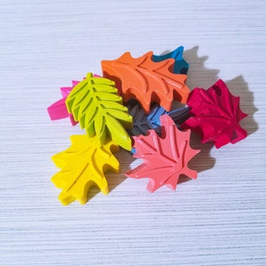 May include: A collection of colorful leaf-shaped crayons in various shades of red, orange, yellow, green, and blue. The crayons are arranged in a pile on a white surface.