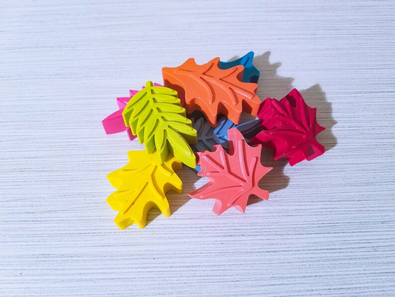 May include: A collection of colorful leaf-shaped crayons in various shades of red, orange, yellow, green, and blue. The crayons are arranged in a pile on a white surface.