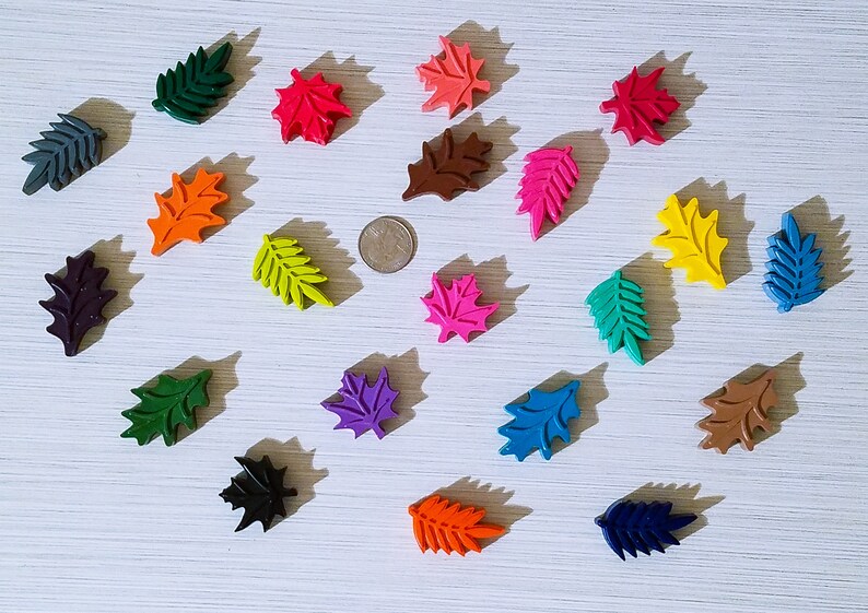 May include: Assortment of 17 leaf-shaped crayons in various colors, including green, red, orange, yellow, pink, purple, blue, brown, and black. The crayons are arranged in a circle on a white surface.