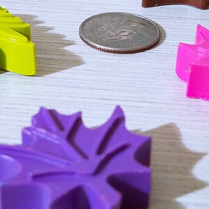 May include: Four colorful leaf-shaped objects made of wax, in shades of green, purple, and pink, are arranged on a white wooden surface. A silver coin is also visible in the image.