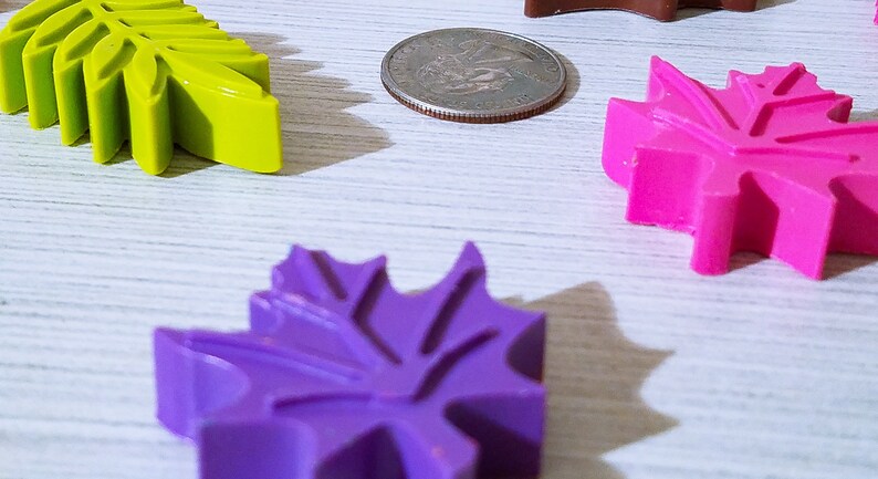 May include: Four colorful leaf-shaped objects made of wax, in shades of green, purple, and pink, are arranged on a white wooden surface. A silver coin is also visible in the image.