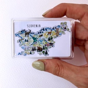 Può includere: Un magnete rettangolare con una mappa ad acquerello della Slovenia, con illustrazioni di animali e alberi. La parola "SLOVENIA" è stampata in alto. Il magnete è racchiuso in un supporto di plastica trasparente.