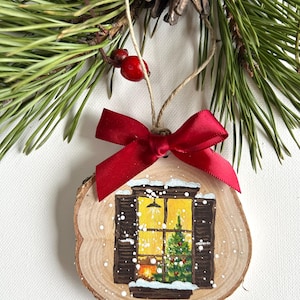 May include: A wooden Christmas ornament with a painted scene of a window with a lit fireplace and a Christmas tree inside. The window is covered in snow and the ornament is decorated with a red bow.