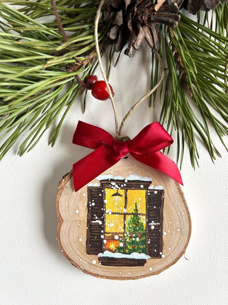 May include: A wooden Christmas ornament with a painted scene of a window with a lit fireplace and a Christmas tree inside. The window is covered in snow and the ornament is decorated with a red bow.