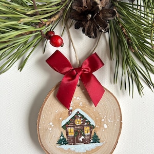 May include: A wooden Christmas ornament with a painted image of a small cabin in a snowy landscape. The ornament is decorated with a red bow and white paint splatters.