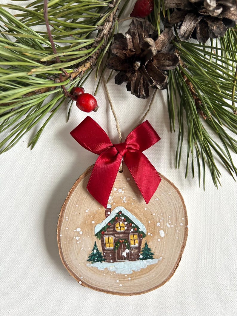 May include: A wooden Christmas ornament with a painted image of a small cabin in a snowy landscape. The ornament is decorated with a red bow and white paint splatters.
