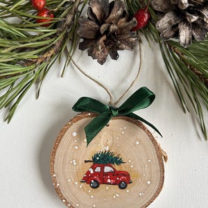May include: A wooden Christmas ornament with a red car carrying a small green Christmas tree on top. The ornament is painted with white dots to resemble snow.