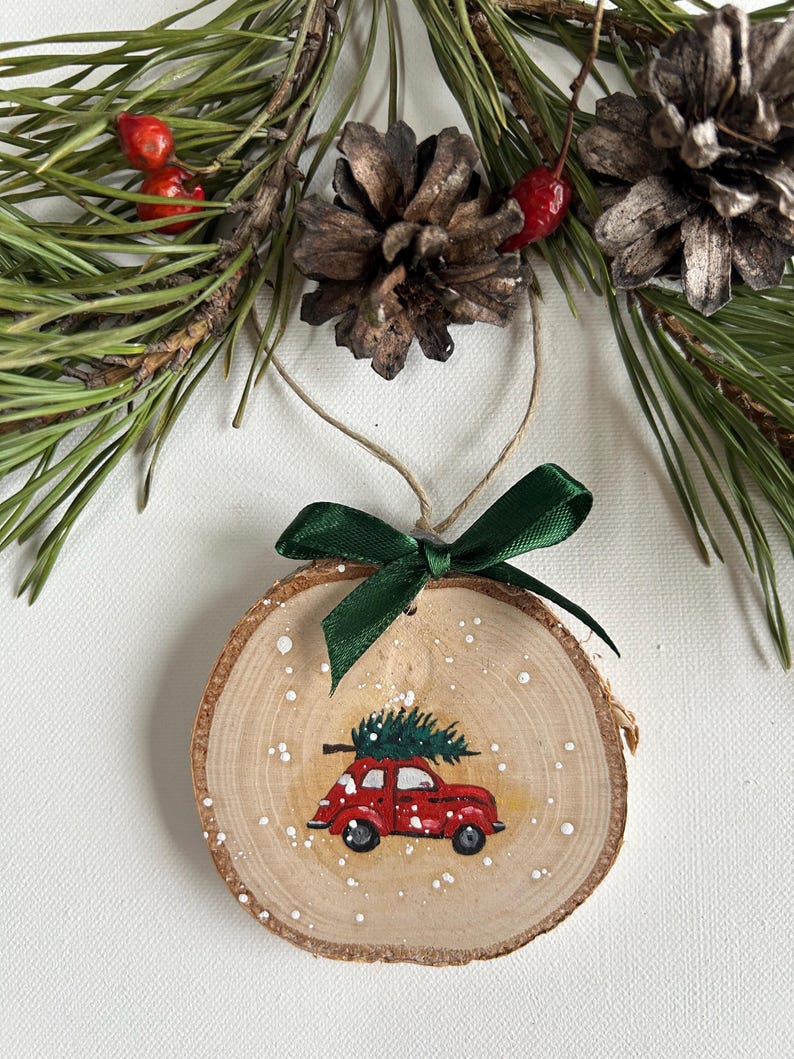 May include: A wooden Christmas ornament with a red car carrying a small green Christmas tree on top. The ornament is painted with white dots to resemble snow.