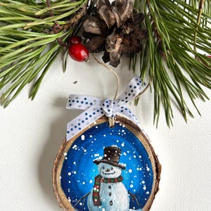 May include: A hand-painted wooden ornament featuring a snowman with a black top hat, red and green scarf, and a blue background with white snowflakes.