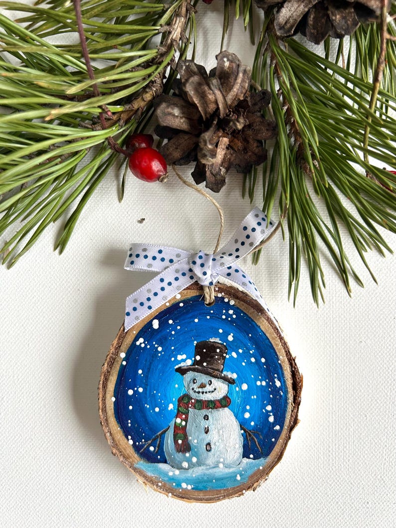 May include: A hand-painted wooden ornament featuring a snowman with a black top hat, red and green scarf, and a blue background with white snowflakes.