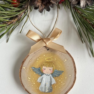 May include: A wooden Christmas ornament featuring a hand-painted angel with a halo, wings, and a white dress. The angel is painted on a round slice of wood with a gold background and gold glitter accents.