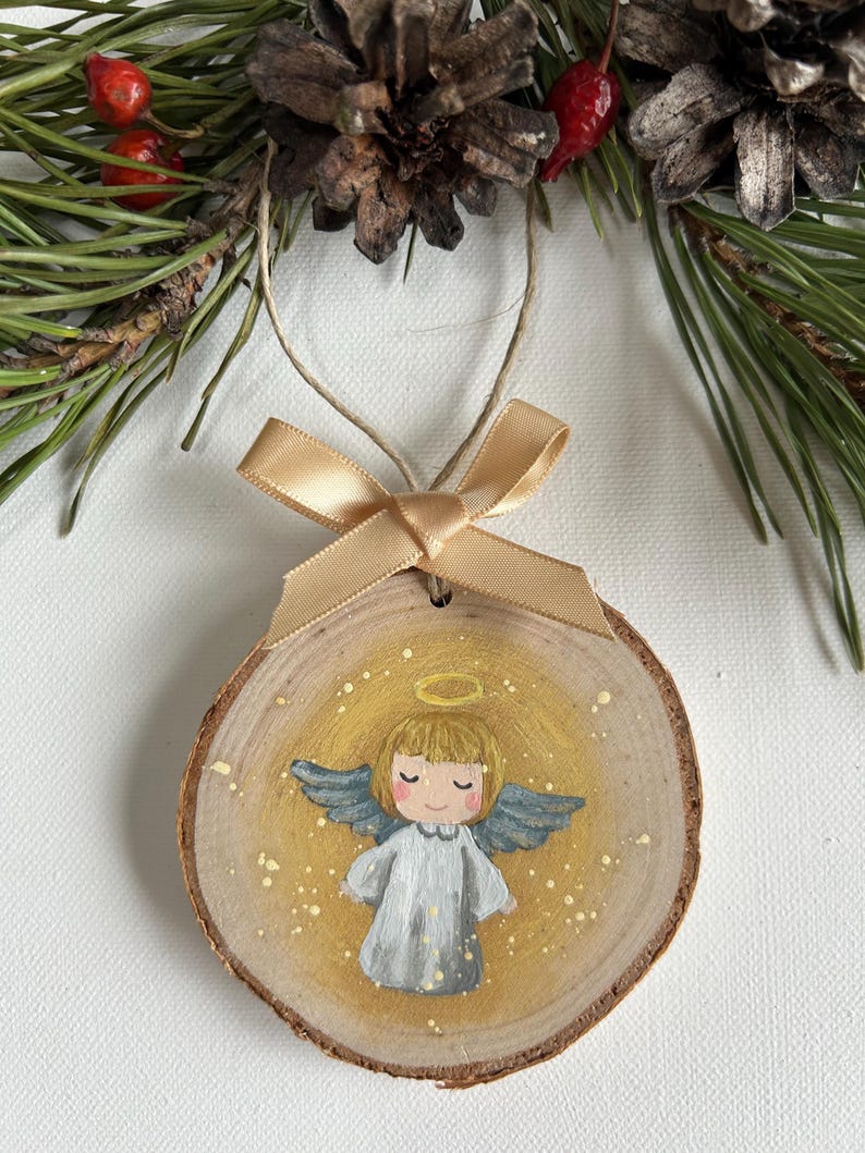 May include: A wooden Christmas ornament featuring a hand-painted angel with a halo, wings, and a white dress. The angel is painted on a round slice of wood with a gold background and gold glitter accents.