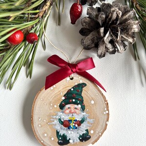 May include: A hand-painted wooden ornament featuring a gnome holding a gift. The gnome is wearing a green hat with red accents and a green coat. The ornament is decorated with white paint to resemble snow.