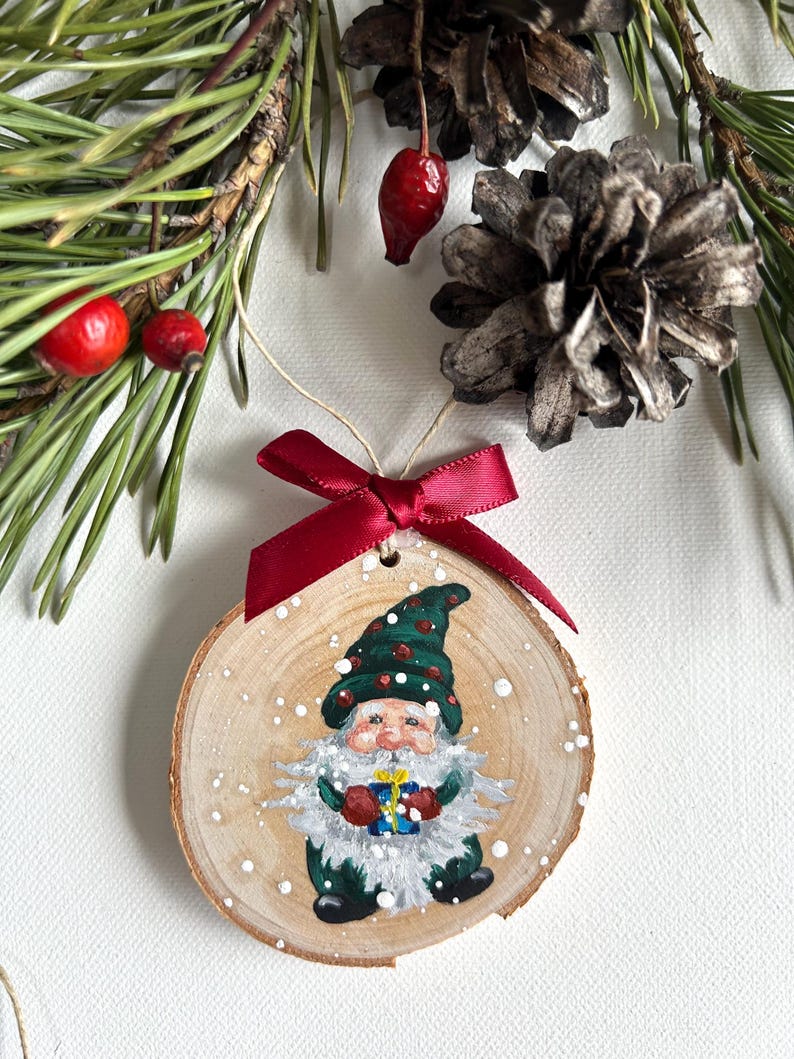 May include: A hand-painted wooden ornament featuring a gnome holding a gift. The gnome is wearing a green hat with red accents and a green coat. The ornament is decorated with white paint to resemble snow.