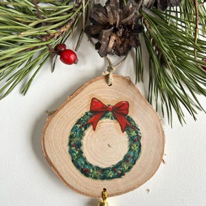 May include: A wooden Christmas ornament with a painted green wreath and a red bow. The ornament has a gold bell attached to the bottom.