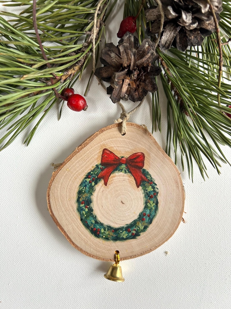 May include: A wooden Christmas ornament with a painted green wreath and a red bow. The ornament has a gold bell attached to the bottom.