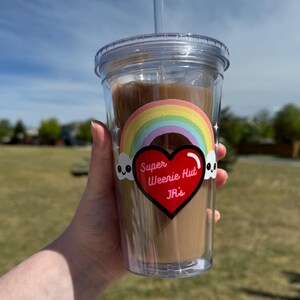 May include: Clear plastic tumbler with a straw, filled with a brown drink. The tumbler features a rainbow sticker with the text "Super Weenie Hut Jr's" and skull graphics. The background is a sunny day with a blue sky and green grass.