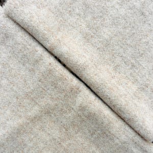 May include: A piece of fabric with a soft, fuzzy texture in a light beige and cream color palette. The material features a subtle, mottled appearance with small flecks of tan distributed throughout the surface, showing a diagonal fold.