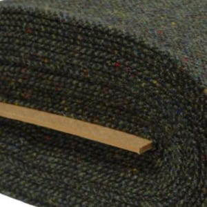 May include: A roll of dark olive green tweed fabric featuring a textured weave with small, multicolored flecks of red, blue, and yellow throughout. The fabric is wrapped around a flat, light brown cardboard core, showing a dense, woven pattern.