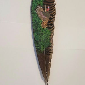 May include: A large, brown and tan feather with a painted scene of a turkey in a forest. The turkey is brown, purple, and red. The forest is green. The feather has a white tip and the artist's signature.