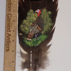 May include: A hand-painted turkey feather with a detailed painting of a wild turkey in a forest setting. The feather is approximately 7 inches long and has a brown and white color scheme. The artist's signature, "W. Scheide - 21", is visible on the feather.