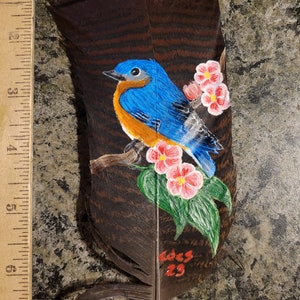 May include: A hand-painted bluebird perched on a branch with pink flowers, painted on a brown feather. The feather has the date "Dec 23" written on it.