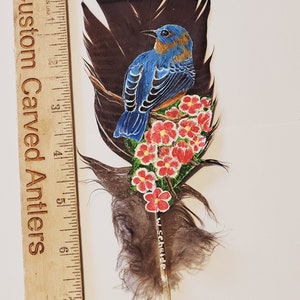 May include: A hand-painted bird on a feather with red flowers. The bird is blue with a brown breast and is perched on a branch. The feather is brown and has a white tip. The flowers are red with green leaves. The artist's signature is visible at the bottom of the feather.