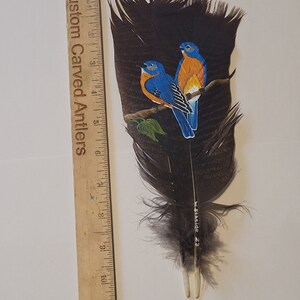 May include: A hand-painted feather with two bluebirds perched on a branch. The birds are painted in shades of blue, orange, and yellow. The feather is brown and has a white quill. The artist's signature, "w.schilde 23", is visible on the feather.