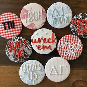 May include: A collection of nine circular fabric buttons featuring embroidered text and patterns. Designs include red and white gingham with TTU, white with TECH, blue floral with ΔΓ mom, black lace with wreck 'em, black lace with guns up, white with wreck 'em, red and white gingham with wreck 'em, blue floral with guns up, and white with ΔΓ.