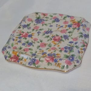 Peut inclure: Plat carré en céramique avec un motif floral. Le plat présente un fond blanc avec des roses roses, des fleurs bleues et des feuilles vertes. Les bords sont bordés d'or. Une pièce décorative pour servir ou exposer.