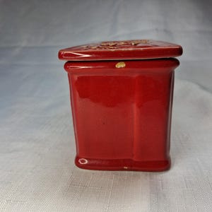 Puede incluir: Una peque&ntilde;a caja de cer&aacute;mica roja con tapa a juego. La caja tiene forma rectangular con lados ligeramente inclinados. La tapa tiene un dise&ntilde;o decorativo. La caja probablemente se utiliza para guardar objetos peque&ntilde;os.