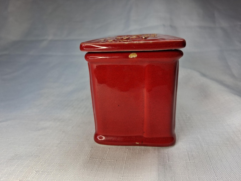Puede incluir: Una peque&ntilde;a caja de cer&aacute;mica roja con tapa a juego. La caja tiene forma rectangular con lados ligeramente inclinados. La tapa tiene un dise&ntilde;o decorativo. La caja probablemente se utiliza para guardar objetos peque&ntilde;os.