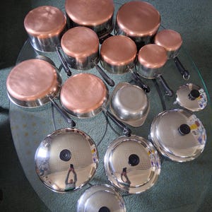 Pode incluir: Um conjunto de panelas e frigideiras com fundo de cobre, laterais de aço inoxidável e cabos pretos. As tampas são de cobre e aço inoxidável. Os utensílios de cozinha estão dispostos sobre uma mesa de vidro.