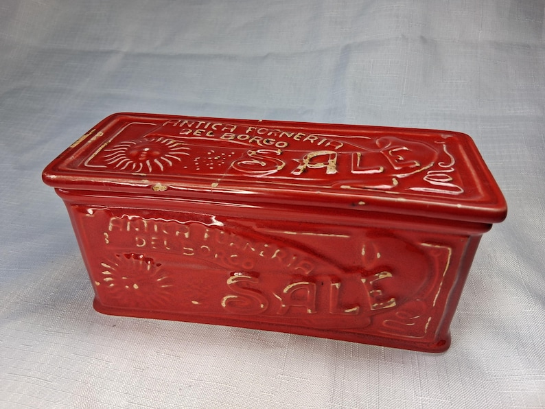Puede incluir: Un recipiente rectangular de cer&aacute;mica roja con tapa. El recipiente tiene texto en relieve que dice "ANTICA FORNERIA DEL BORGO SALE". La palabra "SALE" es prominente. El dise&ntilde;o incluye motivos de rayos de sol y florales.