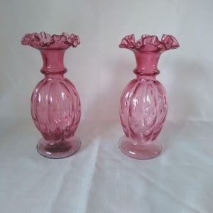 Pode incluir: Dois vasos de vidro rosa com bordas franzidas. Os vasos têm um corpo bulboso com nervuras verticais e uma base alargada. O vidro é translúcido e a cor é rosa intenso.