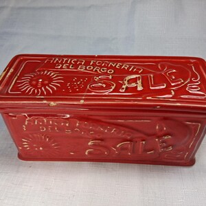 Puede incluir: Una caja rectangular de cer&aacute;mica roja con tapa. La caja presenta texto blanco en relieve que dice "ANTICA FORNERIA DEL BORGO SALE" y un dise&ntilde;o de sol. La caja es probablemente para almacenamiento o exhibici&oacute;n.